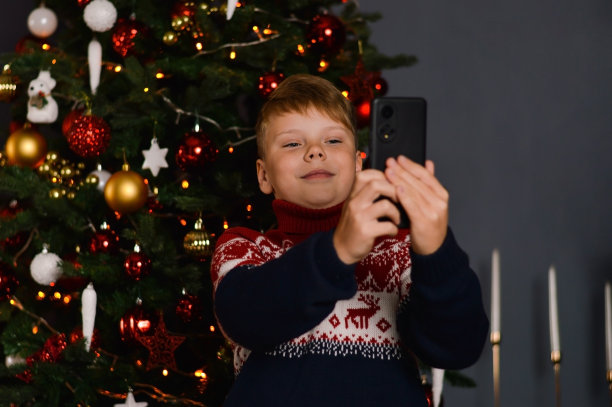 平安夜，男孩微笑着用手机拍自己的照片。圣诞树上有暗红色的灯作为背景。图片下载
