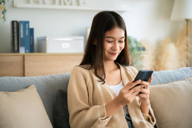 快乐的年轻亚洲女子放松在舒适的沙发上在家里发短信在手机上，微笑的女孩使用手机聊天，浏览无线互联网上的小工具，从家里网上购物图片下载