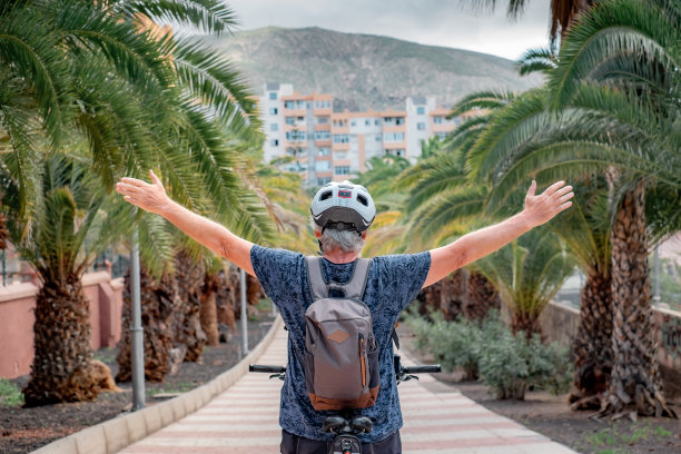 城市公园里戴着头盔的骑自行车男子的背影，他张开双臂，骑着他的电动自行车奔跑。健康的生活方式和可持续的出行理念图片下载