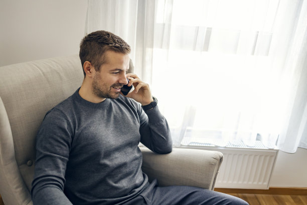 一个年轻人坐在家里舒适的椅子上打电话。图片下载