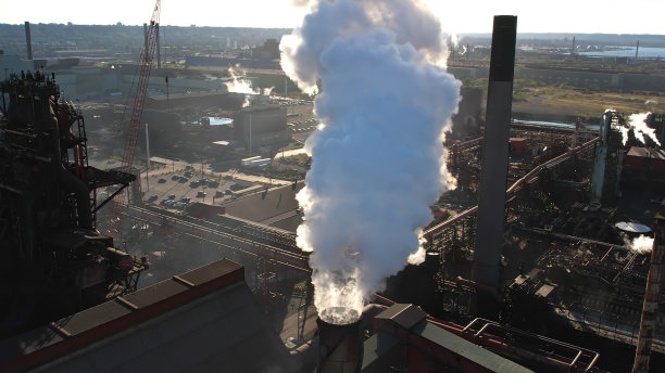 钢铁和采矿工厂的高管道与白色的大烟囱。高空烟囱管与灰色烟雾从生产设施的鸟瞰图。烟雾移动，阻挡夕阳，灯光闪烁图片下载