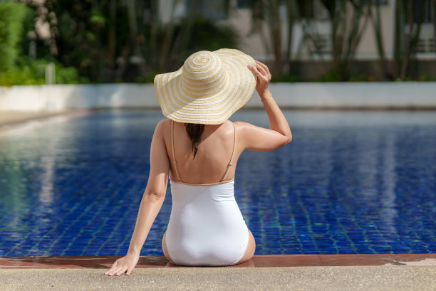 年轻美丽的亚洲女子在白色泳装，戴着大帽子坐在蓝色户外游泳池的边缘在阳光灿烂的日子。图片下载