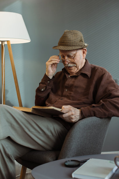 一位老人独自坐在客厅的椅子上，戴着眼镜看书。图片下载