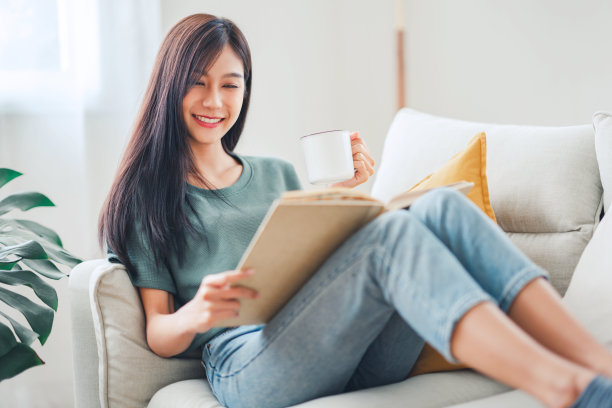 年轻的亚洲女子坐在窗前的现代沙发上，在客厅里看书，在家喝咖啡或茶图片下载