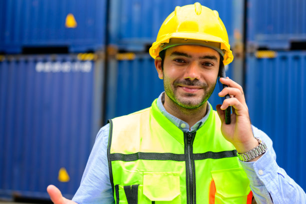 微笑的肖像英俊的男性产业工人工头在黄色安全帽和安全背心说话在手机上控制货物仓库在货物集装箱码头图片下载