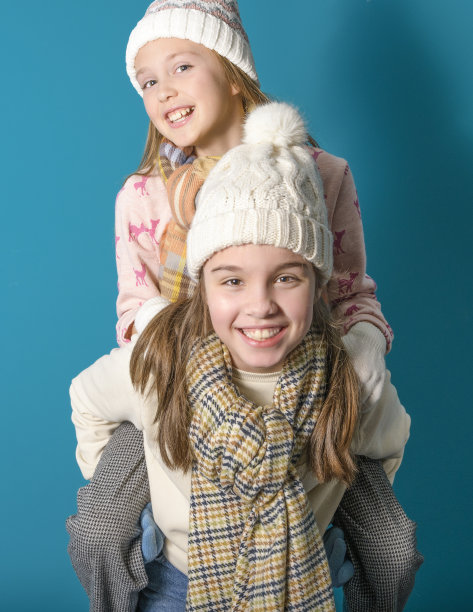 两个姐妹或十几岁的朋友玩跳蛙舞。冬天的主题和衣服。工作室的照片。毛衣，围巾，手套。脸上洋溢着喜悦。垂直在蓝色背景上图片下载