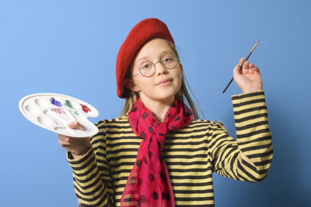 年轻的艺术家。一个女孩戴着鲜红色的贝雷帽，在蓝色背景上拿着一把刷子。特写肖像图片下载