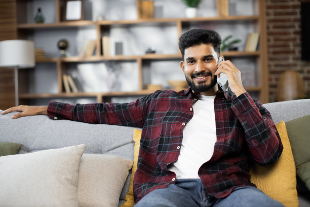 快乐的年轻胡须男子坐在舒适的沙发上，手里拿着现代智能手机打电话图片下载