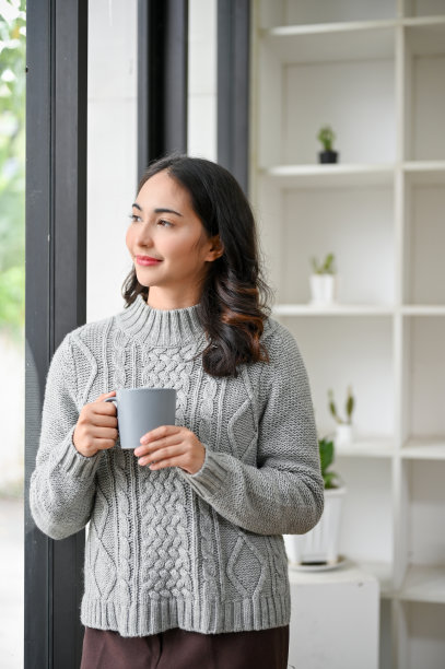 穿着舒适的针织毛衣的亚洲女人站在窗前，喝着咖啡图片下载