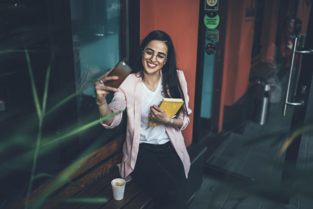 快乐的女作家诗人和教科书，使用手机前置摄像头在休息时点击自拍图片，快乐的格鲁吉亚妇女戴着眼镜在人行道上休闲拍摄视频图片下载