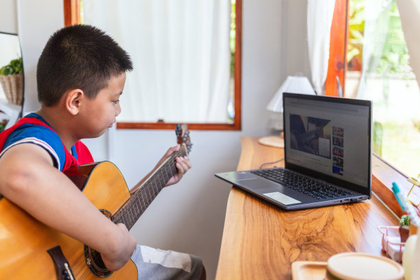 一个男孩看着笔记本电脑，准备在家练习弹吉他的故事。男孩们在网上上古典吉他课。图片下载