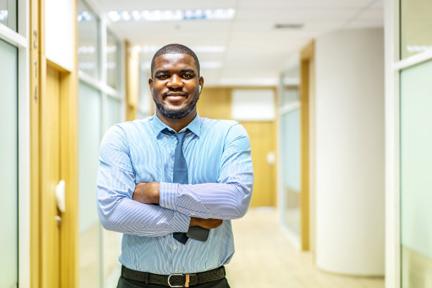微笑成熟的非洲裔美国行政商人在西装看着相机，而双臂交叉摆姿势在现代走廊办公室的肖像图片下载