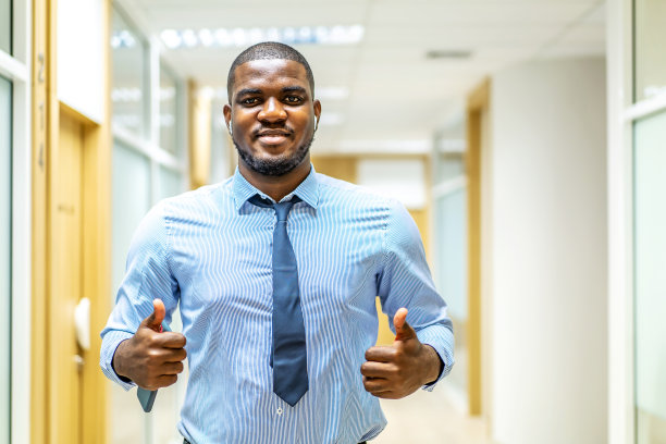 微笑的肖像成熟的非洲裔美国行政商人在西装看着相机，同时显示大拇指在现代走廊办公室摆姿势，同意签字，笑得很开心图片下载
