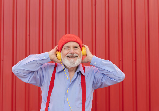 有趣的微笑退休男子触摸耳机看着相机孤立在红色背景的照片图片下载