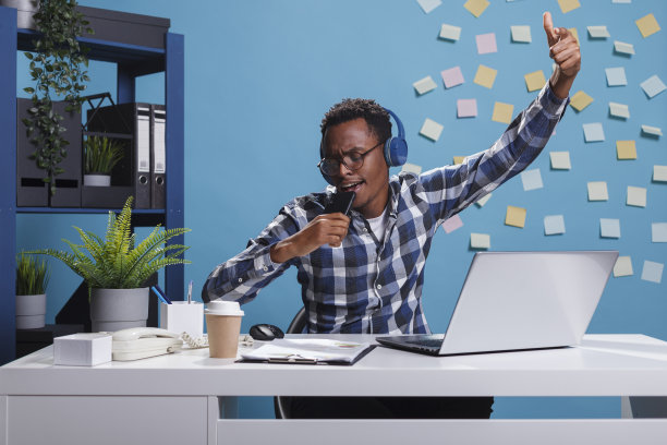 兴奋开朗的非裔美国上班族戴着无线耳机，坐在办公桌前唱卡拉ok。图片下载