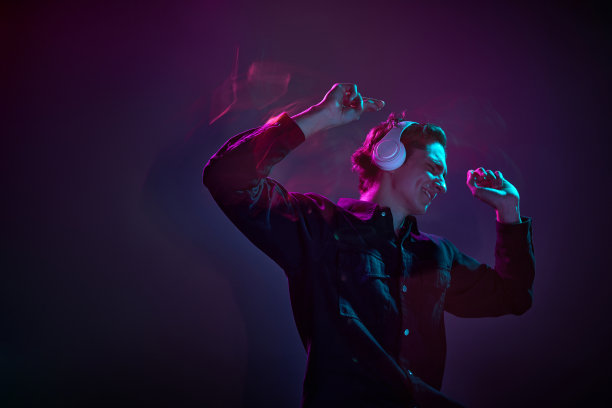 年轻男子的肖像在休闲黑色衬衫，跳舞的耳机隔离在渐变色深紫色背景在霓虹灯混合光。人类情感的概念，面部表情。图片下载