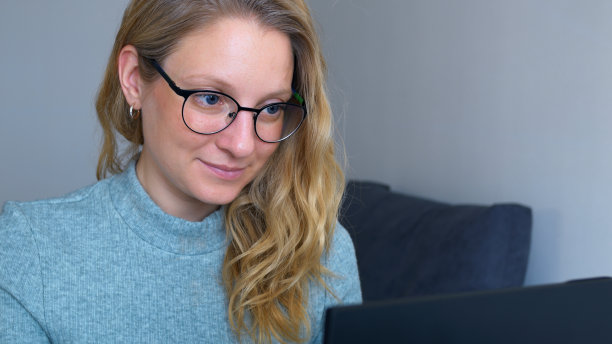 戴眼镜的年轻成功女性积极地在办公室的电脑前工作，并享受她的工作。女秘书正在她的工作场所用笔记本电脑打字。女人把她的金发藏在耳朵后面。图片下载