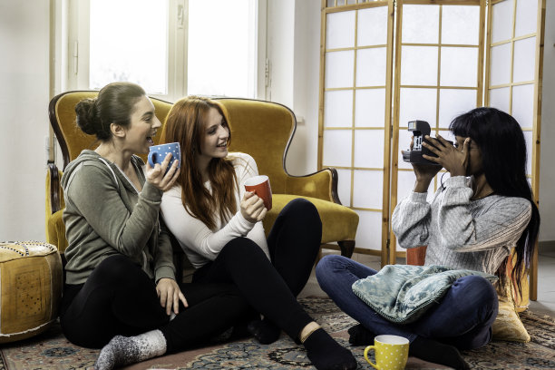 快乐的多元文化女性朋友在家里一起享受自由时光-快乐的年轻女性在客厅喝咖啡或茶，用老式相机拍照图片下载