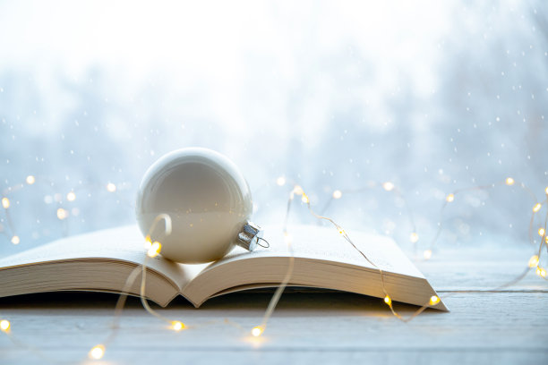 白色的圣诞装饰物在打开的纸质书上，白色的桌子上有仙女灯，背景是雪域森林景观。冬季或圣诞节阅读概念。景深浅。图片下载