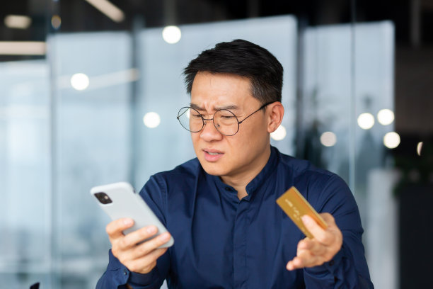 受骗的网上买家，办公室里的亚洲商人试图在网上商店进行银行交易和购买，男子手持银行信用卡和手机不高兴图片下载
