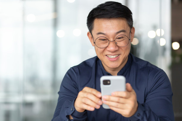 特写照片:一个快乐和微笑的亚洲男人从电话中阅读快乐的消息，一个穿着衬衫和眼镜的商人微笑和欣喜，一个男人在办公室里靠窗户工作图片下载