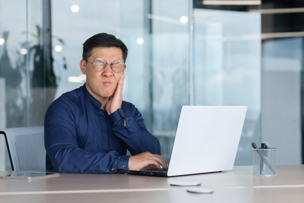 亚洲人在办公室生病了，商人牙痛，男人坐在大楼里的办公室里用笔记本电脑工作，在窗户边的衬衫让老板心烦意乱图片下载