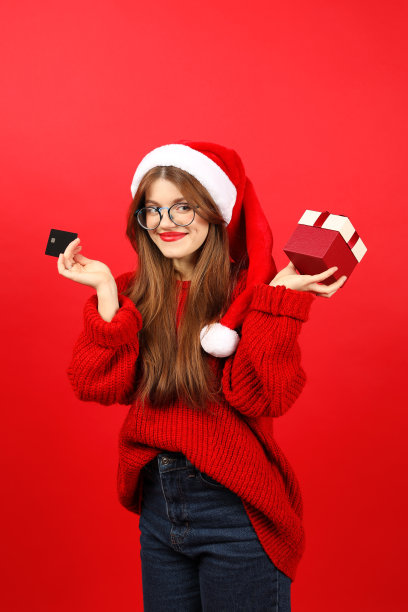 用支付卡买圣诞礼物。一名年轻女子戴着红色背景的圣诞老人帽子，手里拿着一个惊喜。圣诞节打折促销。图片下载