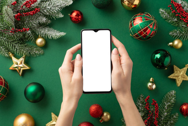 圣诞节的概念。第一人称俯视图照片，女性手拿智能手机，红色，金色，绿色装饰物，星星饰品，槲寄生浆果和冷杉树枝，孤立的绿色背景与空白图片下载