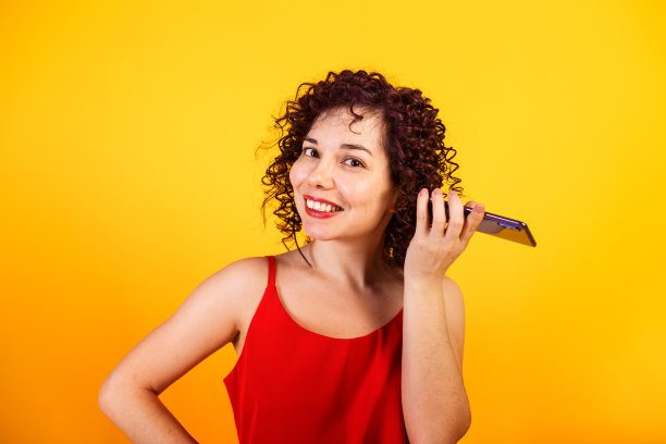 可爱的微笑卷发女孩正在打电话。一位年轻女士正在听语音信息。工作室女性肖像在明亮的黄色背景。图片下载