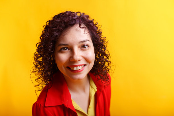 工作室拍摄的正面和微笑的女孩。穿着鲜红衬衫和黄色t恤的年轻卷发女子。黄色背景隔离。图片下载