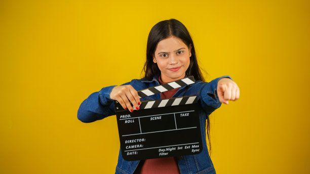 美丽的年轻亚裔印度女子站着拿着隔板，食指指向相机，隔板用于电影制作，隔离在彩色背景的工作室肖像图片下载