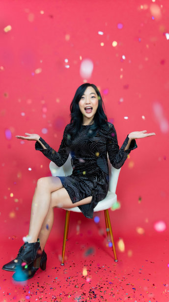 假日，新年和派对概念:美丽的年轻女子与礼盒穿着晚礼服坐在五彩纸屑雨在红色背景。快乐的时间。图片下载