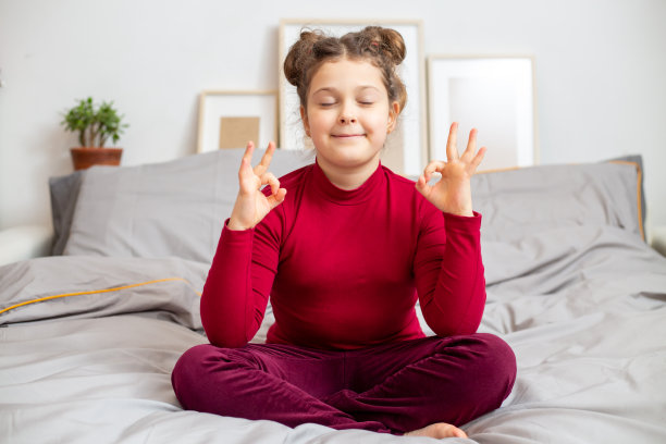 快乐平静的小白种人女孩10在一个灰色的床上冥想在家里的酒红色t恤。孩子在家中通过瑜伽放松身心，以冥想为健康生活的锻炼方式。健康的生活方式。家庭生活图片下载