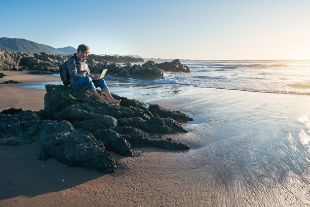 一个成熟的拉丁数字游牧民，在日落时分坐在海滩的岩石上用笔记本电脑工作图片下载
