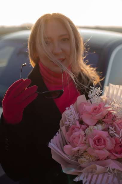 垂直接近时尚的金发女子与墨镜在黑色夹克粉红色玫瑰花束反对黑色汽车图片下载