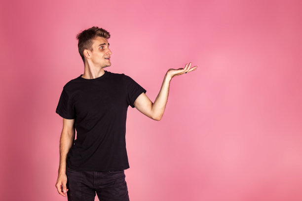 肖像年轻男子在黑色t恤展示产品与手掌在粉红色的背景，看向别处。有人在空地上举手。广告展示产品概念。复制文本空格图片下载