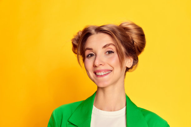 年轻开朗的女孩在明亮的绿色夹克摆姿势的肖像，微笑在黄色的工作室背景。积极的图片下载