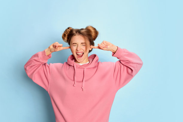 年轻女孩的肖像在粉红色卫衣摆姿势，关闭耳朵烦恼的蓝色工作室背景。刺激图片下载