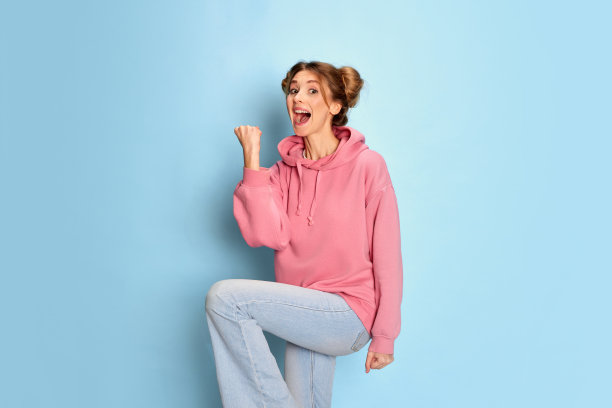 年轻开朗的女孩在粉红色卫衣的肖像与积极的表情在蓝色的工作室背景。成功图片下载