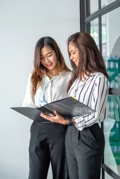 两个年轻漂亮的亚洲女商人站在一起，拿着平板电脑做文书工作，讨论项目财务报告。公司业务协作概念。图片下载