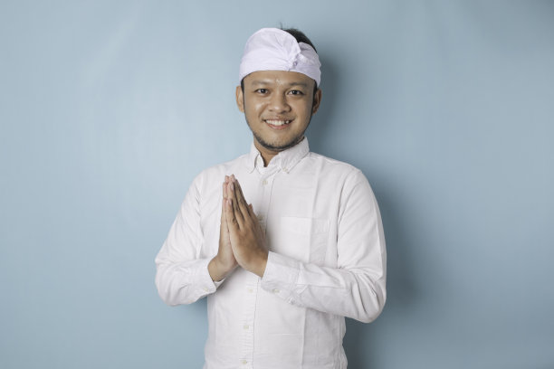 微笑的年轻巴厘男子戴着乌登或传统的发带和白色衬衫，手势问候或合十礼孤立在蓝色背景图片下载