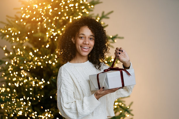 一名非裔美国妇女在房子舒适的客厅的圣诞树旁拿着一份节日礼物图片下载