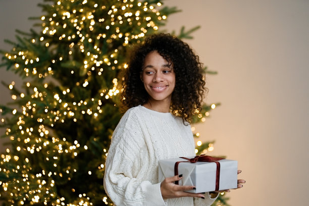一名非裔美国妇女在房子舒适的客厅的圣诞树旁拿着一份节日礼物图片下载