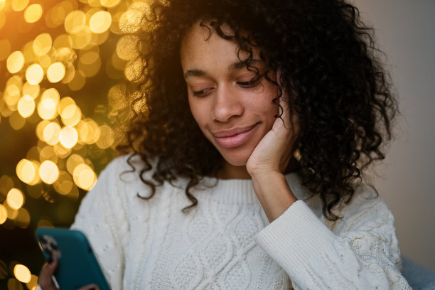 一名非裔美国妇女在舒适的客厅的圣诞树旁用智能手机聊天图片下载