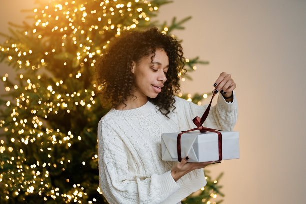 一名非裔美国妇女在房子舒适的客厅的圣诞树旁拿着一份节日礼物图片下载