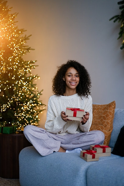 一名非裔美国妇女在房子舒适的客厅的圣诞树旁拿着一份节日礼物图片下载