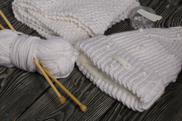 白色针织帽和围巾。女式手工针织服装。旁边有一捆用来编织的白线和针织针。用于装饰容器的珠子。在黑板上。图片下载