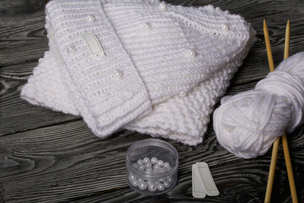 白色针织帽和围巾。女式手工针织服装。旁边有一捆用来编织的白线和针织针。用于装饰容器的珠子。在黑板上。图片下载