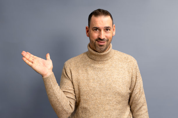 一个留着胡子，穿着棕色高领毛衣的西班牙人，友好地在他的手掌上提供一些东西，孤立在灰色的背景上。图片下载