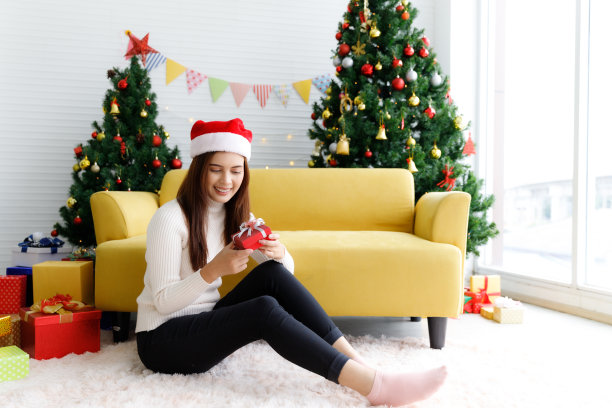 Smiling brunette woman in sweater and Santaâs hat sitting on the floor in living room holding small gift box and rejoices over Christmas tree decorative background图片下载